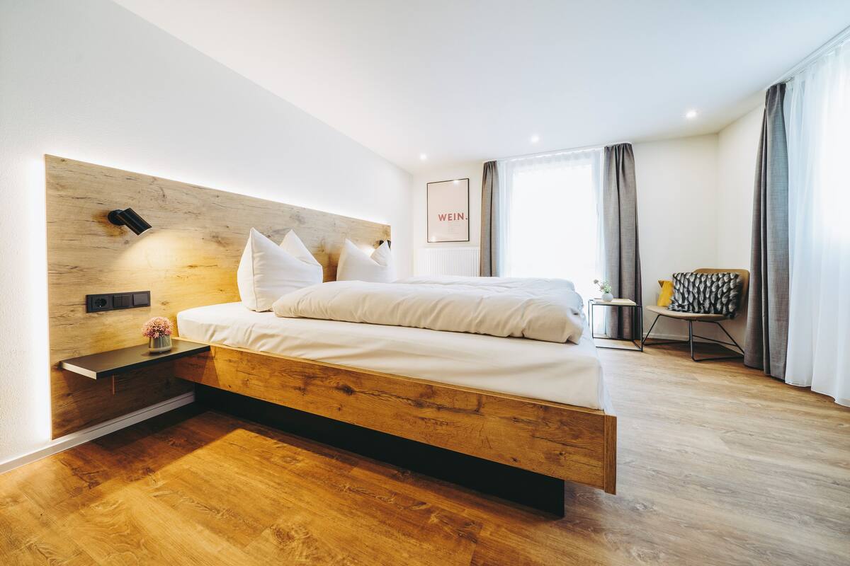 A modern hotel room with a wooden headboard, a bed with white linens, a bedside table with a lamp, and a view of the city through large windows.
