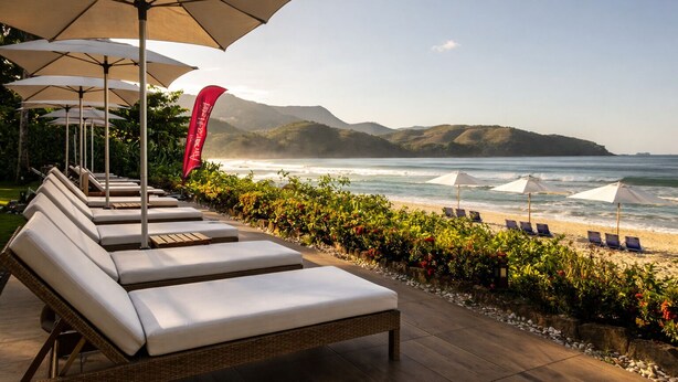 A beachside lounge area with sun loungers and umbrellas, a red banner, and a view of the ocean and hills. - Amora Hotel (São Sebastião)