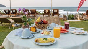 A breakfast setup with a view of the ocean, including chairs, a table with plates, cups, and a fruit basket. - Amora Hotel (São Sebastião)