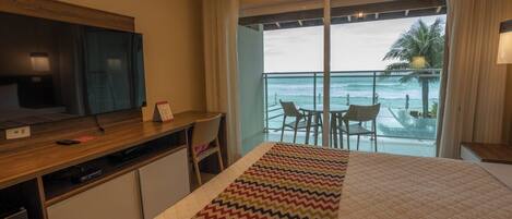Une chambre d’hôtel avec un lit, un bureau avec une télévision, une petite table et un balcon avec des chaises et vue sur l’océan.