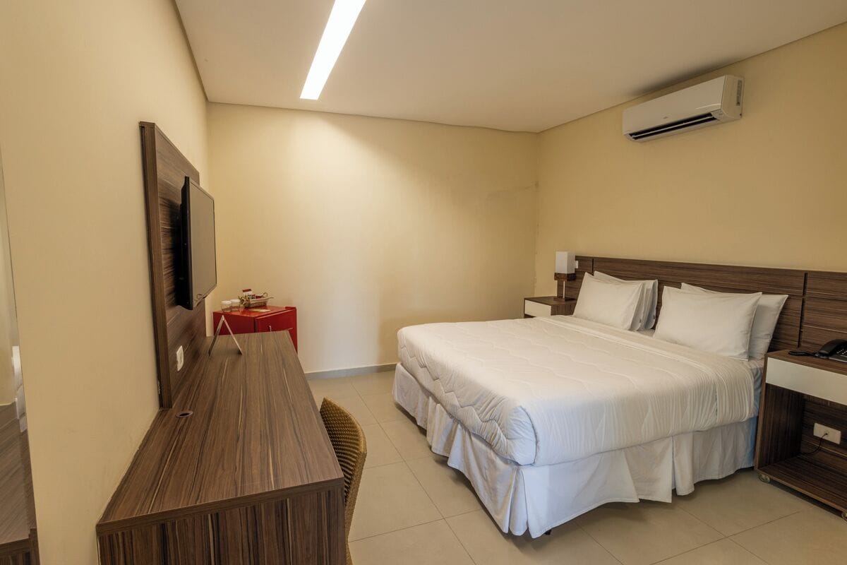 A hotel room with a bed, bedside table, a desk, a chair, an air conditioning unit, and a television.