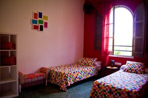 A room with two single beds, a small table, a shelf with a rolled-up item, and a window with red curtains. - Home Backpackers Valencia by Feetup Hostels (Valencia)