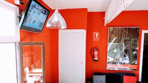 A room with a red wall, a grey sofa, a black and white checkered floor, a television, and a window with a reflection of a person. - Home Backpackers Valencia by Feetup Hostels (Valencia)