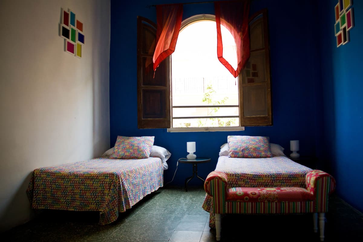 A room with two single beds, a small table, and a window with red curtains.