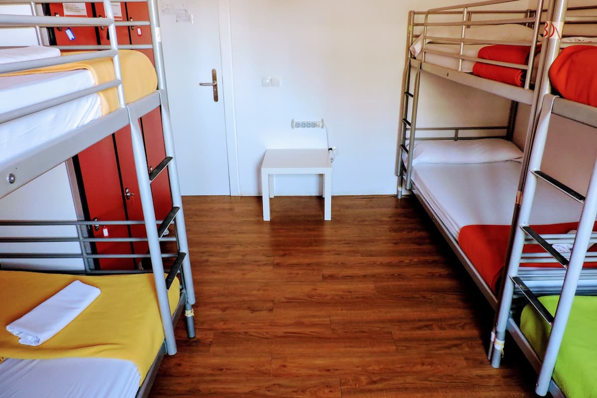 A dormitory room with bunk beds, a small white bench, and a wooden floor.