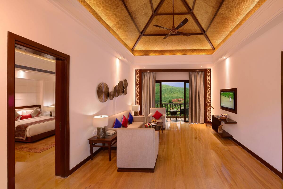 A hotel room with a large bed, a sofa, a TV, and a balcony with a view of the hills.