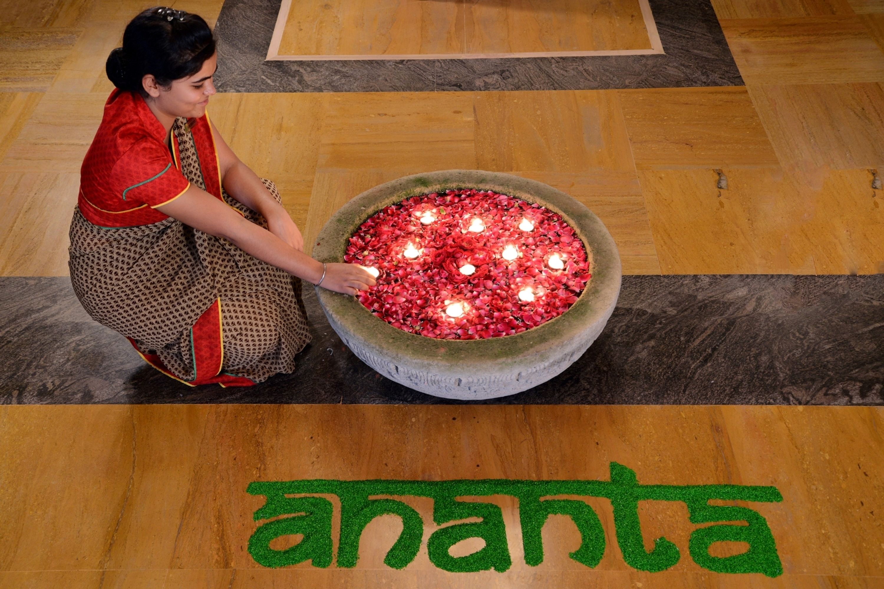 Een vrouw die rode bloemblaadjes in een grote kom rangschikt op een houten vloer, met het woord 'ananta' in het groen.