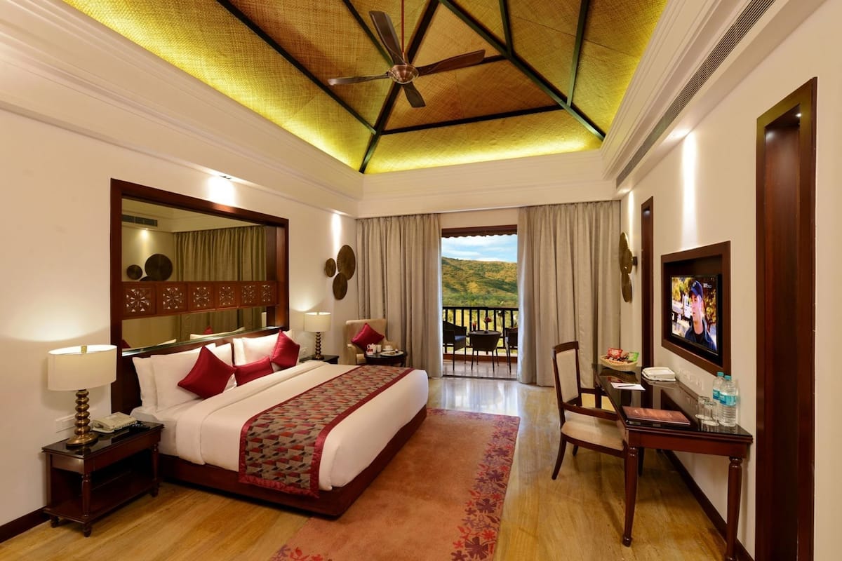 A hotel room with a bed, a desk with a TV, a balcony with a view, and a ceiling fan.