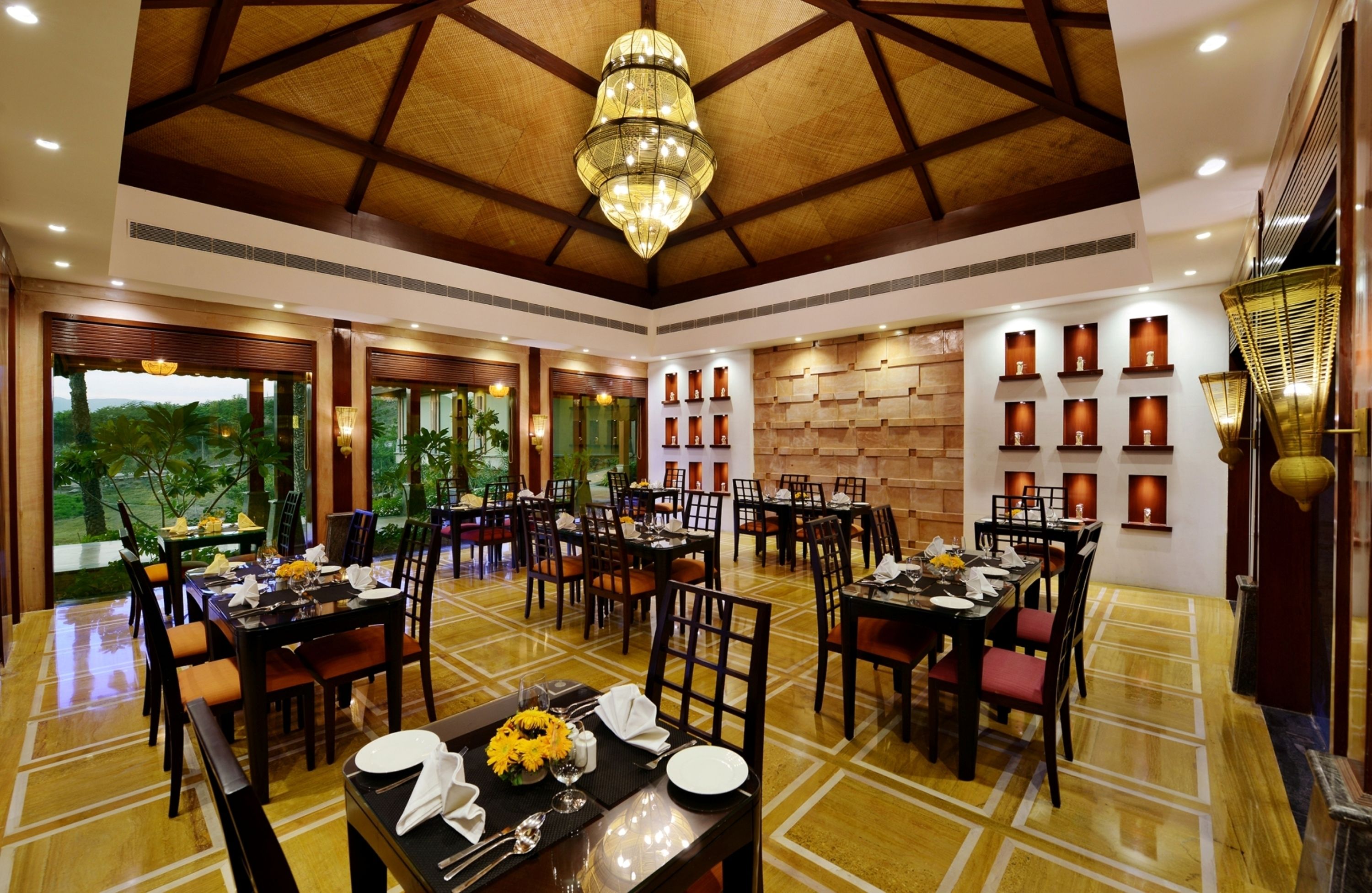 A spacious dining area with multiple tables and chairs, a large chandelier, and a view of greenery outside.