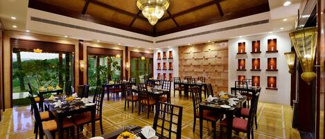 A spacious dining area with multiple tables and chairs, a large chandelier, and a view of greenery outside.
