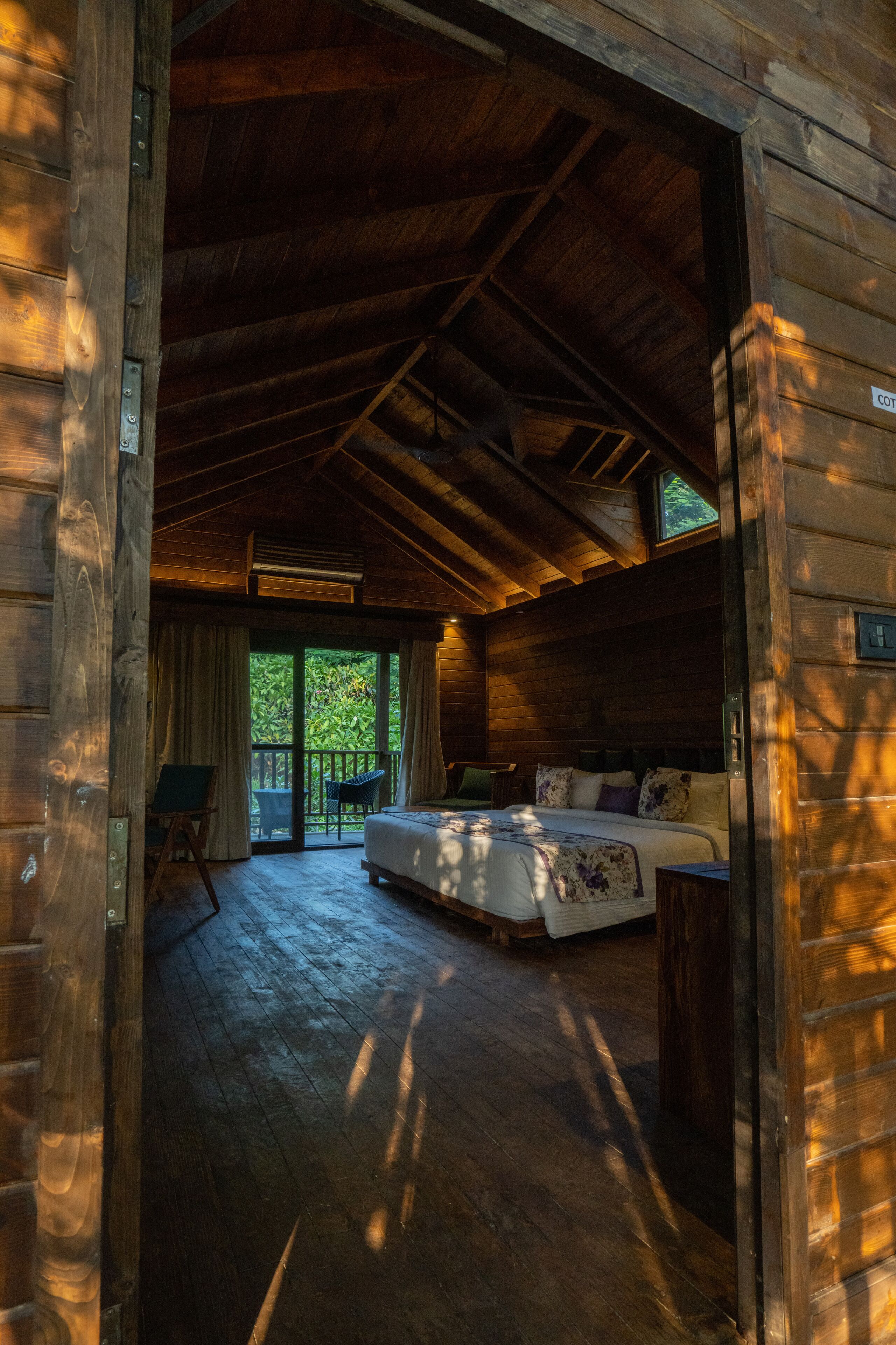 A wooden cabin bedroom with a bed, a desk, and a window overlooking greenery.