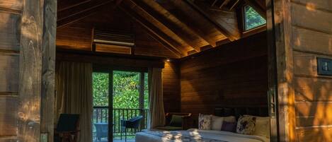 A wooden cabin bedroom with a bed, a desk, and a window overlooking greenery.