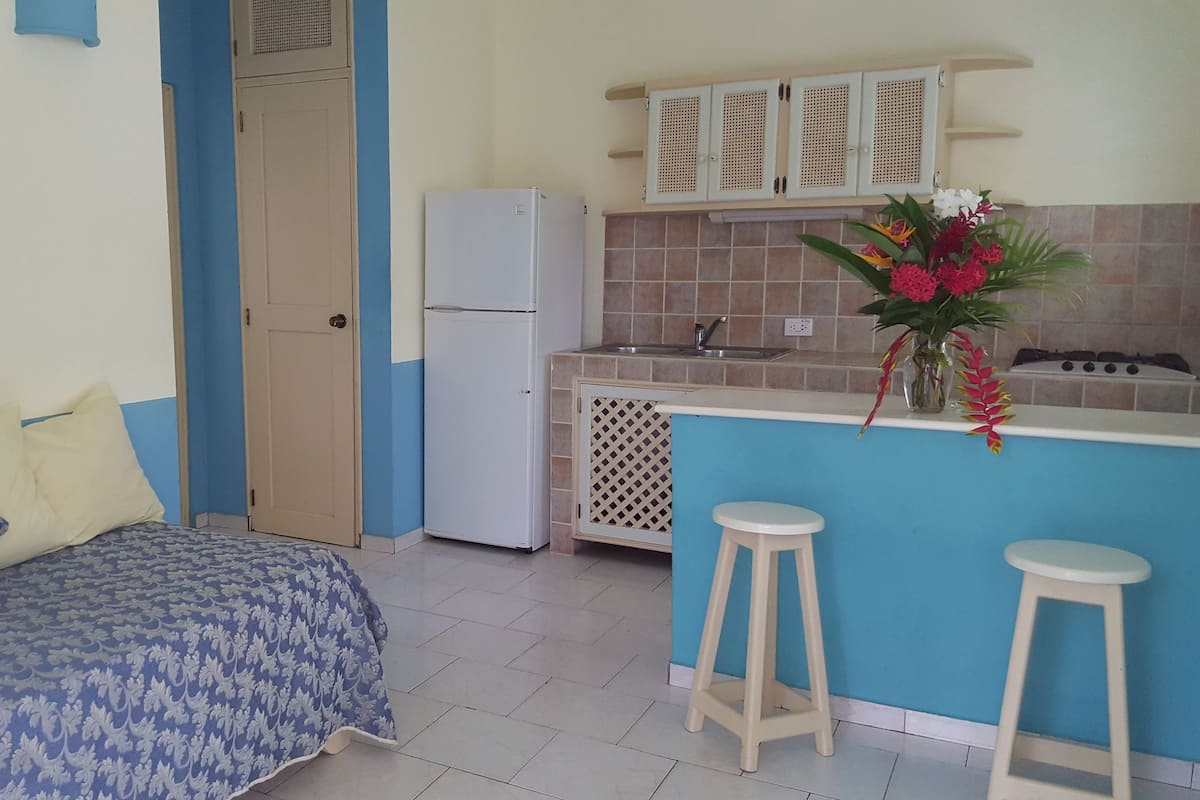 A compact kitchen with a refrigerator, a sink, and a stove. There is a blue sofa with patterned cushions and a small stool.