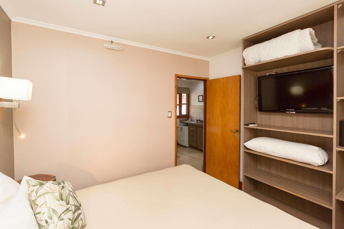 A hotel room with a bed, a television on a wooden shelf, and a kitchenette in the background.