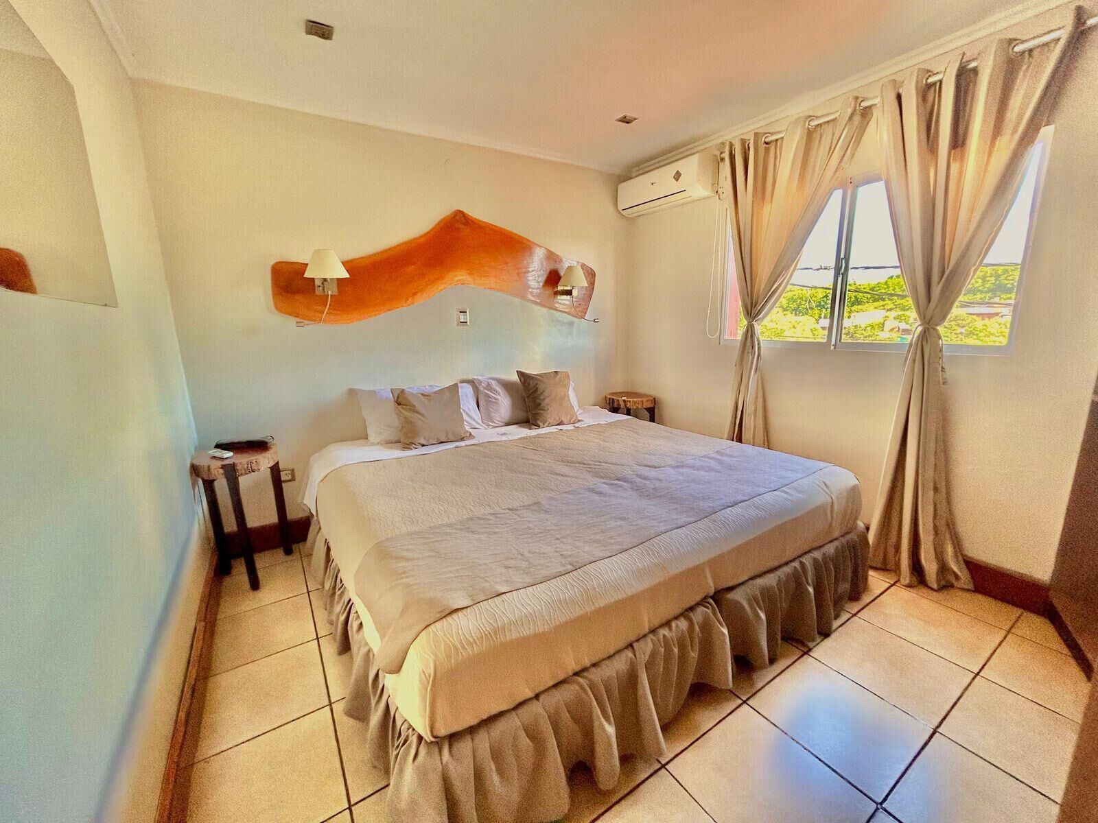 A bedroom with a bed, a nightstand, an air conditioning unit, a window with curtains, and a view of greenery outside.