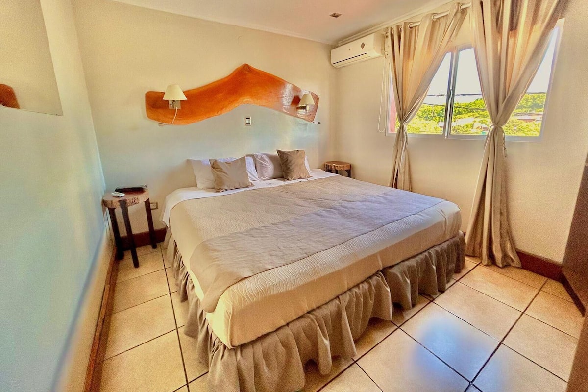 A bedroom with a bed, a nightstand, an air conditioning unit, a window with curtains, and a view of greenery outside.