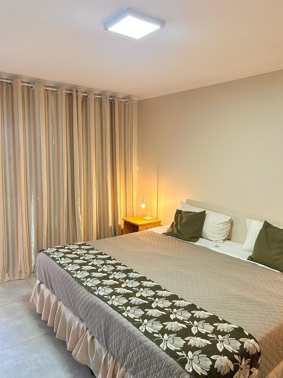 A hotel room with a bed, curtains, a bedside table with a lamp, and a window.