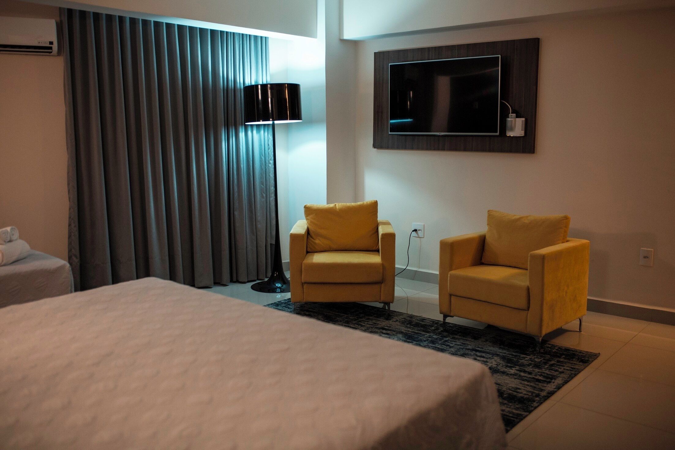 Une chambre d’hôtel avec un grand lit, deux fauteuils jaunes, un téléviseur fixé au mur et une fenêtre avec des rideaux.