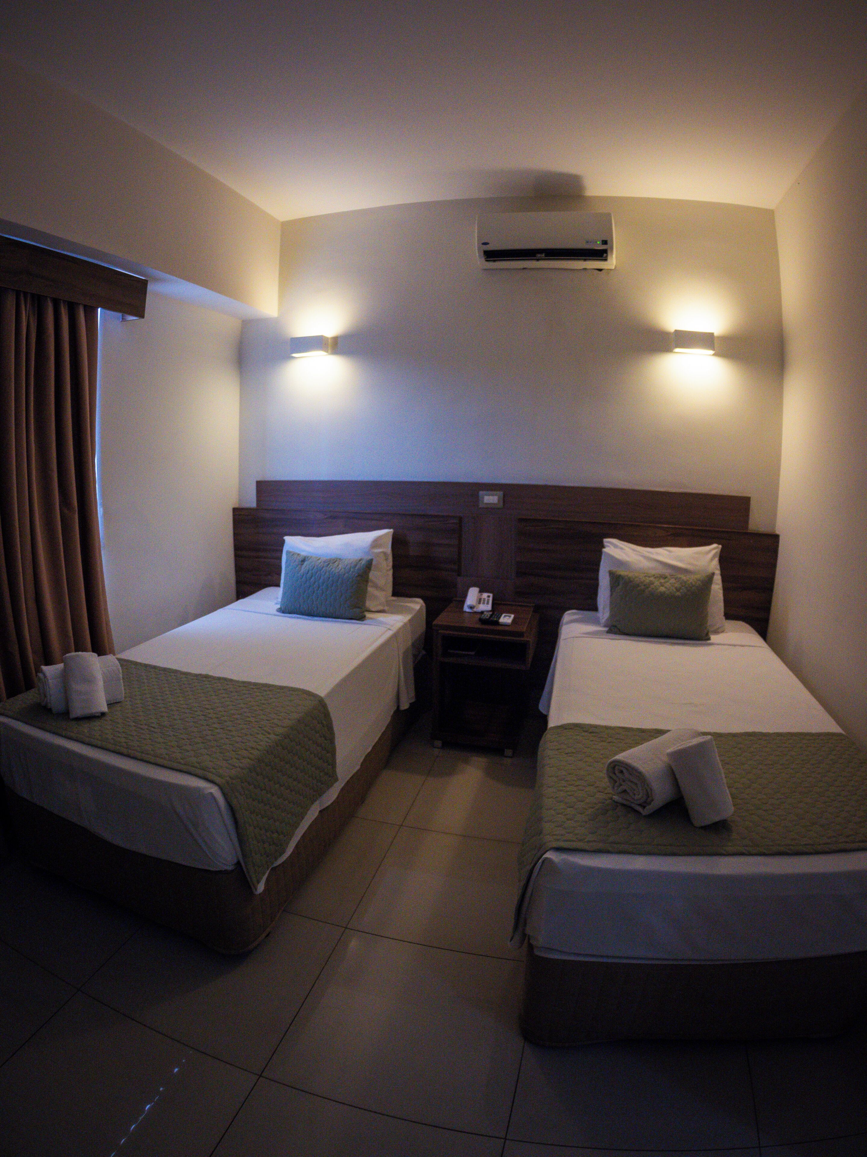 A hotel room with two beds, a nightstand, an air conditioning unit, and a window with curtains.