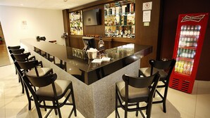 Een bar met een lange toonbank, meerdere stoelen, een televisie, planken met flessen en een rode drankdispenser.