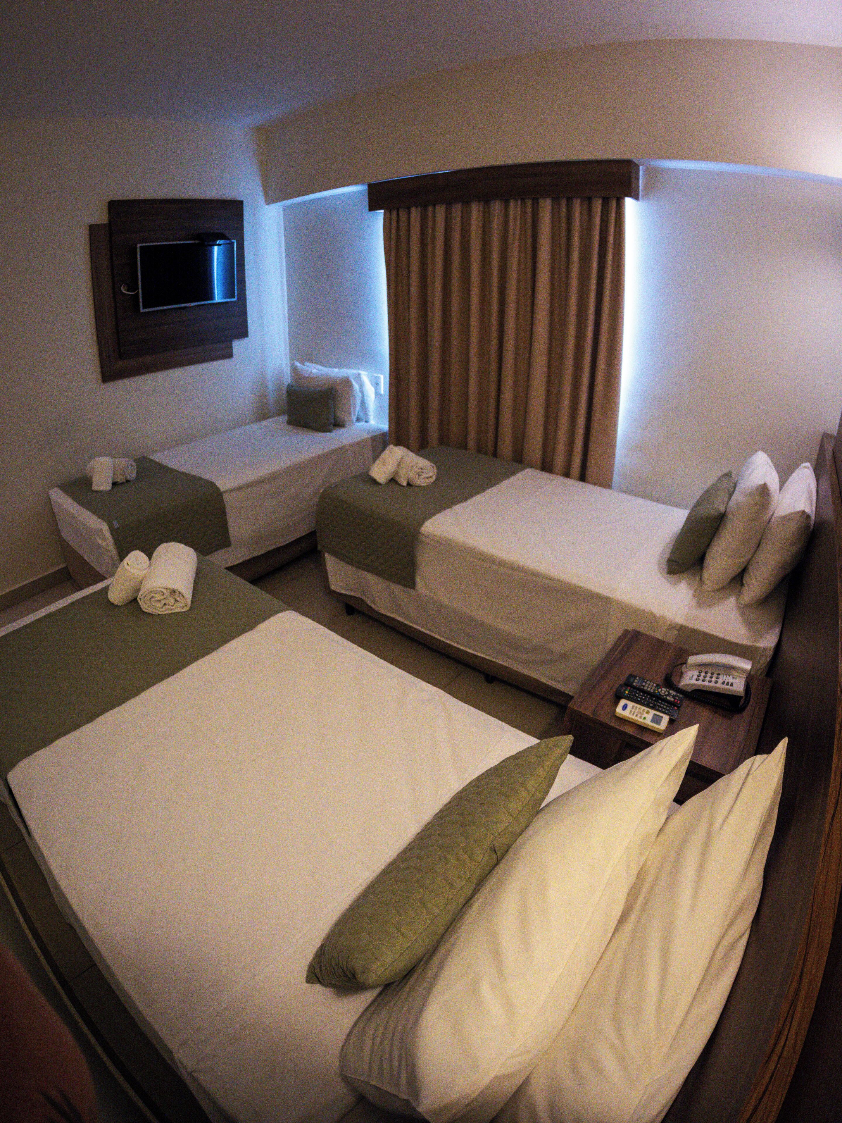Habitación de hotel con dos camas individuales, un televisor montado en la pared, una mesita de noche con teléfono y controles remotos, y una ventana con cortinas.