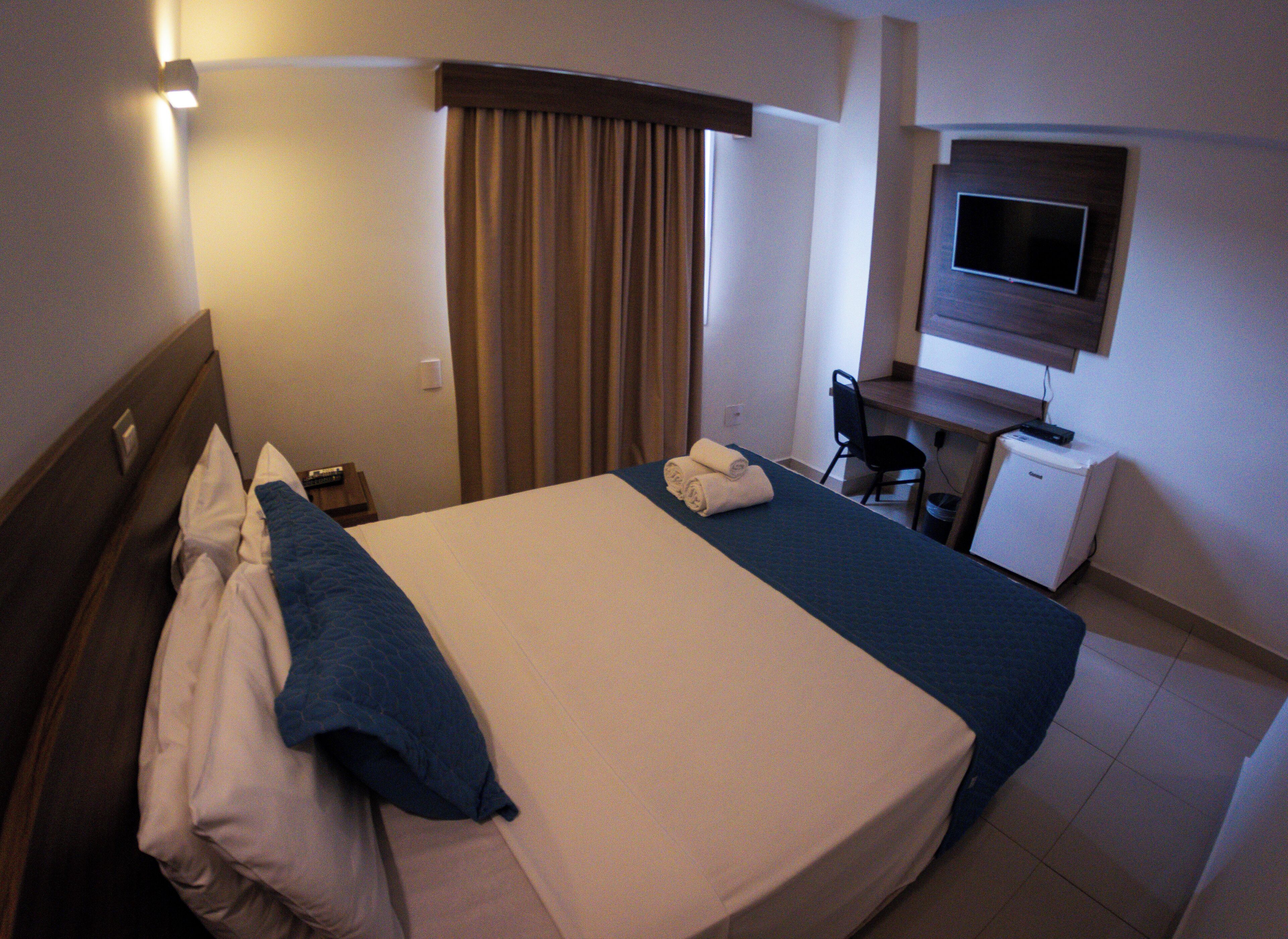 Quarto de hotel com cama, escrivaninha, cadeira e televisão.
