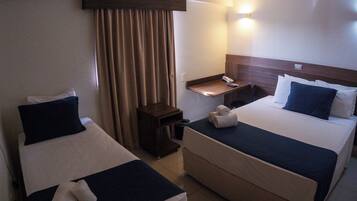 Quarto de hotel com duas camas, uma secretária pequena, uma mesa de cabeceira e uma luminária na parede.