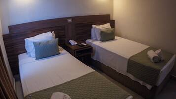 A hotel room with two single beds, each with a green bedspread and white linens. There is a nightstand with a telephone and a remote control. An air conditioning unit is mounted on the wall above the beds.