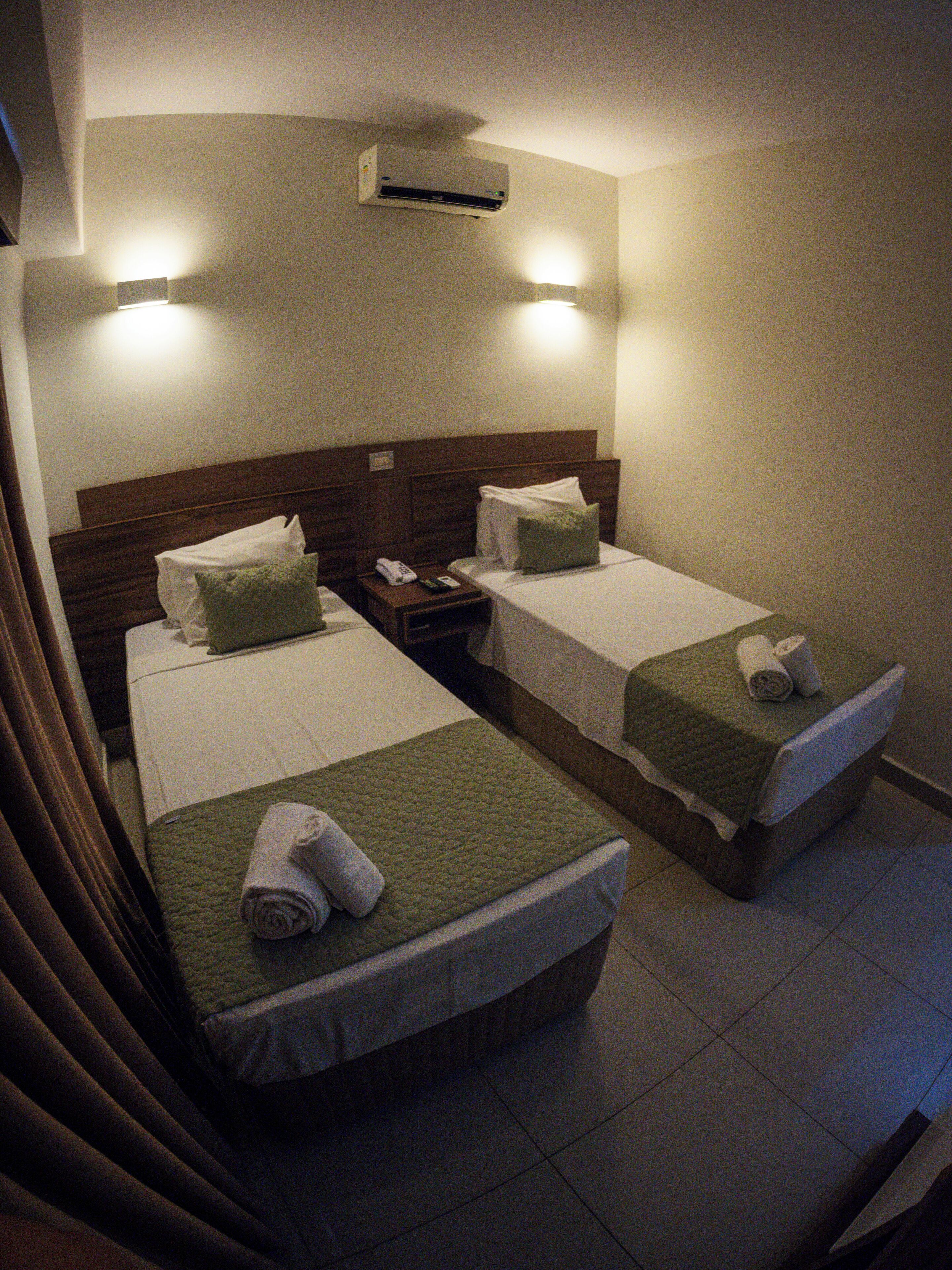 Ein Hotelzimmer mit zwei Betten, jedes mit einer grünen Decke und weißen Laken. Ein Nachttisch mit Telefon und Fernbedienung sowie eine Wandaircondition sind vorhanden.