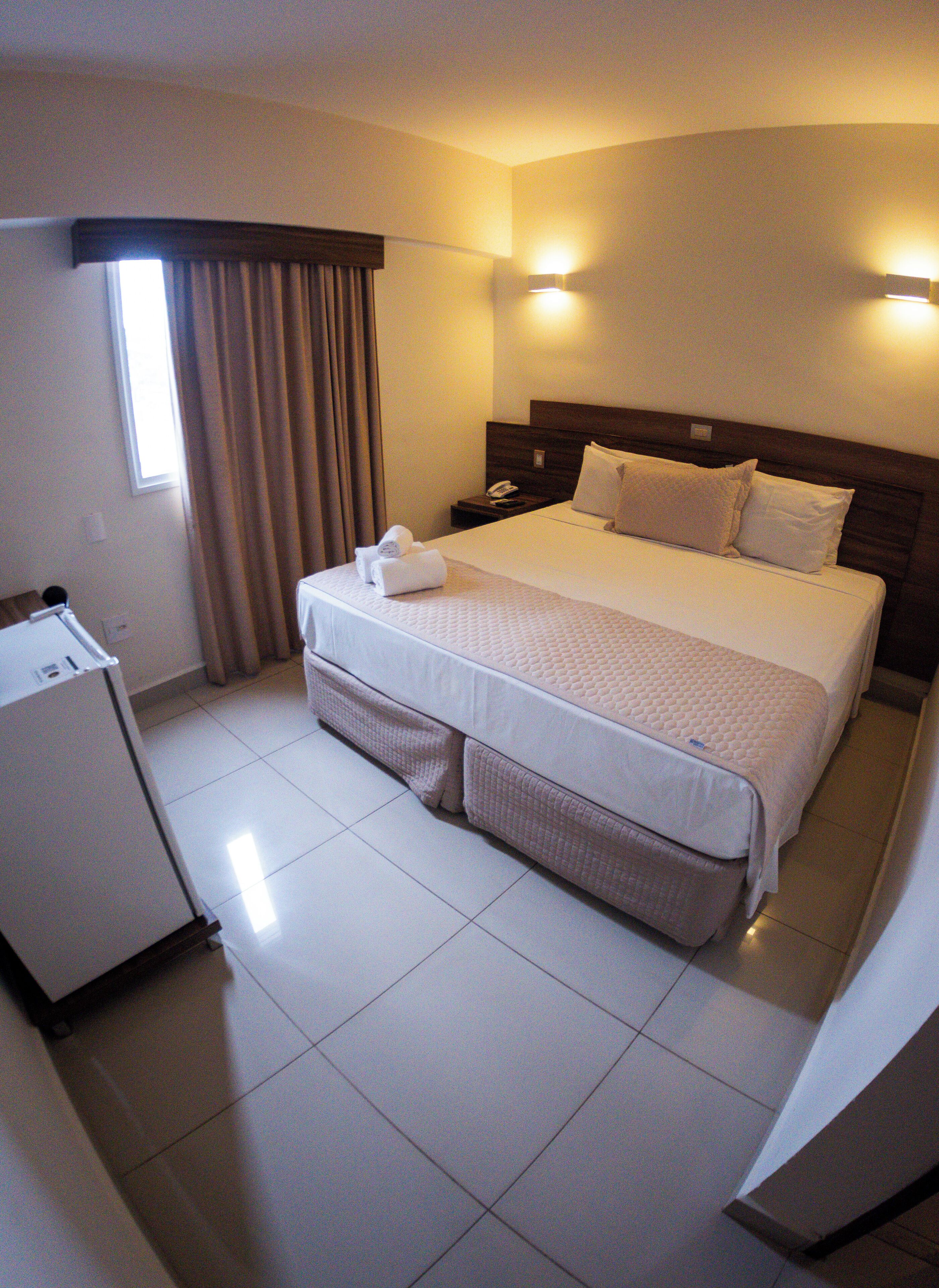 A hotel room with a bed, a bedside table, a lamp, a window with curtains, and a refrigerator.