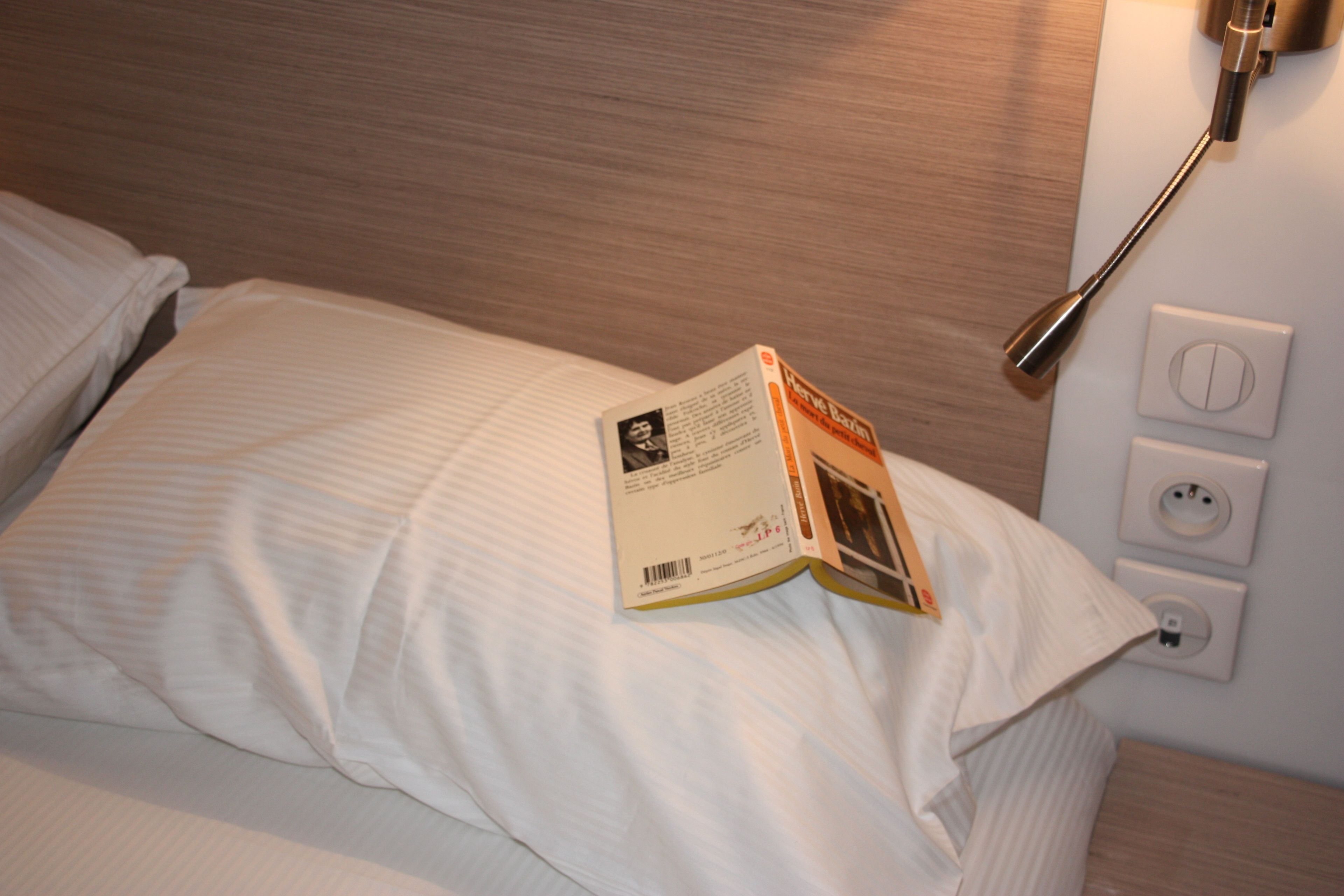 Een netjes opgemaakt bed met een boek erop, een bedlampje en een stopcontact aan de muur.