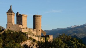 Un château médiéval doté de trois tours, entouré d’une végétation luxuriante et avec des montagnes en toile de fond.