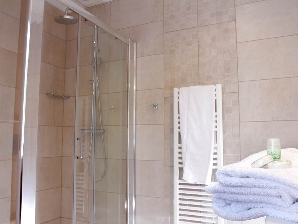 Een douchecabine met glazen wanden, een handdouche, een witte handdoekhouder met een opgevouwen handdoek en een fles lotion op een witte ondergrond.