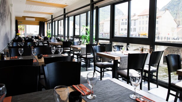 Un restaurant avec des tables dressées pour le service, de grandes fenêtres offrant une vue sur les bâtiments et les montagnes.