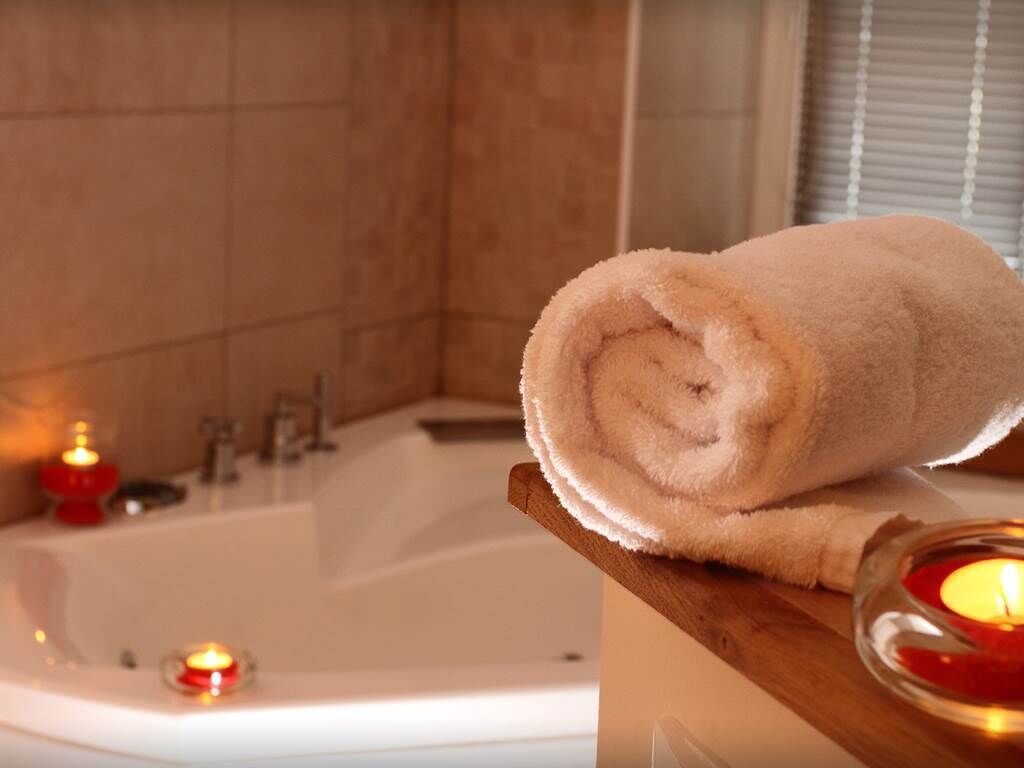 Een badkamer met een badkuip, een opgerolde handdoek op een houten plank en kaarsen.