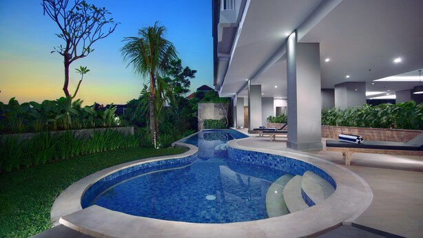 A modern outdoor pool area with a curved design, surrounded by well-maintained greenery and a clear sky. - Hotel Neo Denpasar by ASTON (Denpasar)