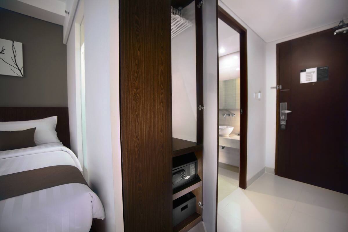 A hotel room with a bed, a wooden wardrobe, a bathroom with a sink and mirror, and a door with a sign.