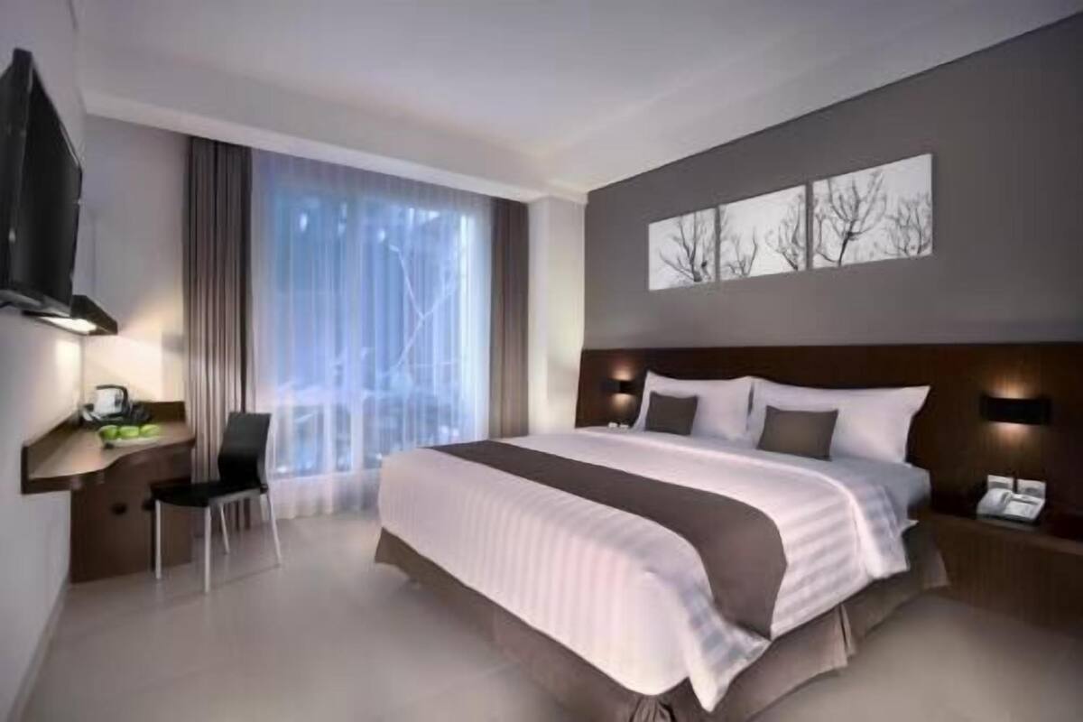 A modern hotel room with a large bed, a desk with a chair, a flat-screen TV, and a large window with sheer curtains.