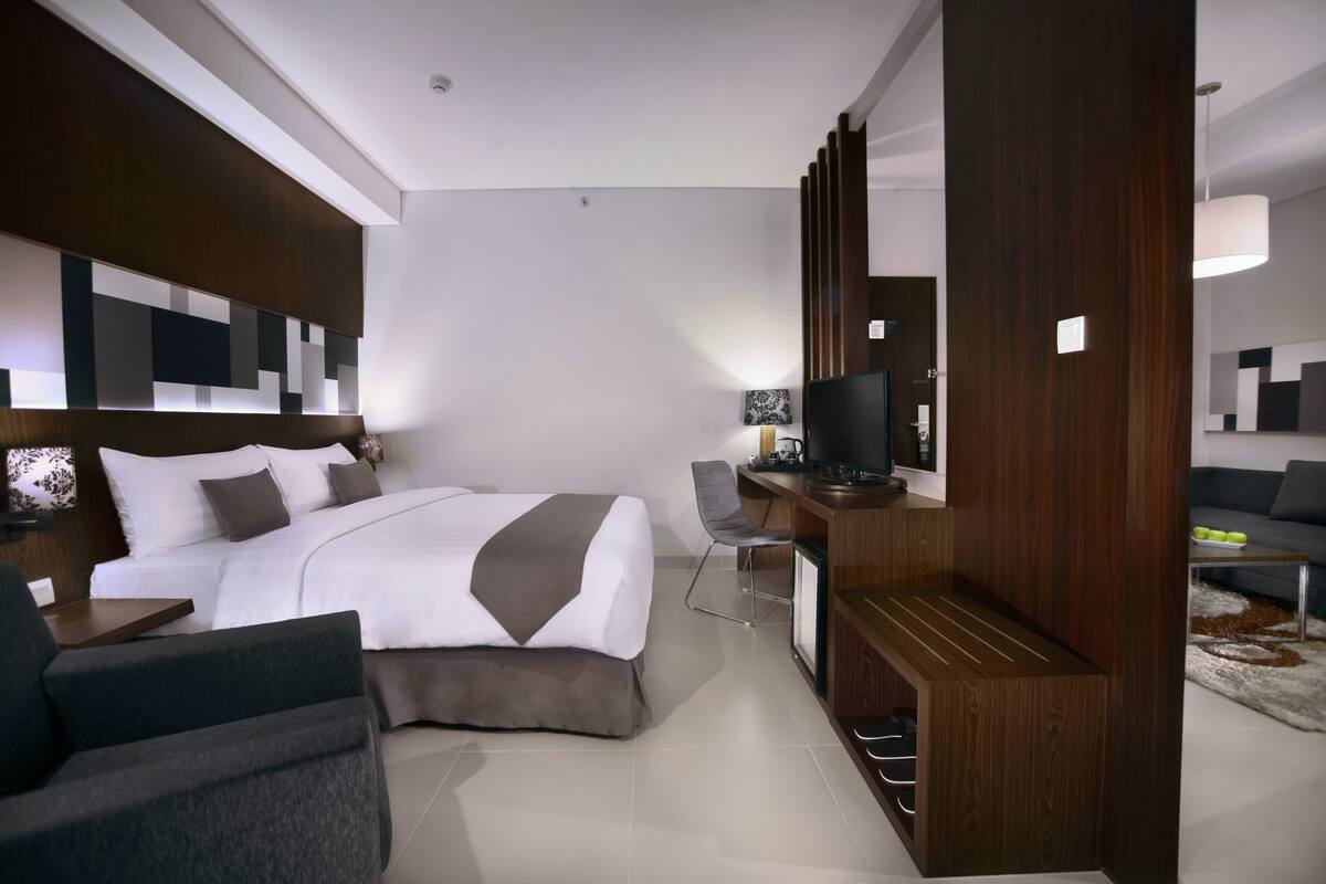 A modern hotel room with a neatly made bed, a desk with a computer, a television, and a sofa.