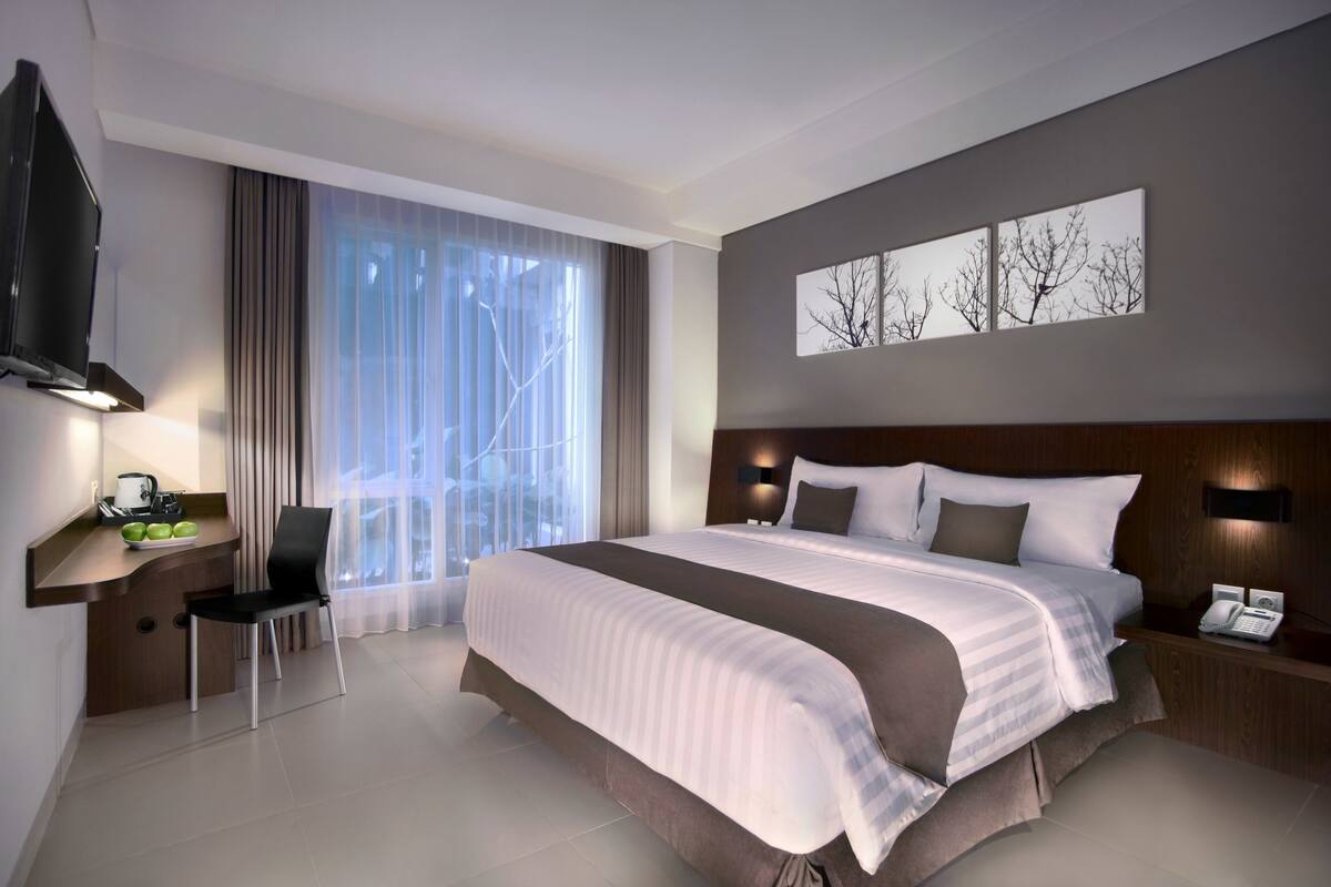 A modern hotel room with a large bed, a desk with a chair, a flat-screen TV, and a large window with curtains.