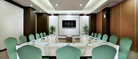 A conference room with a long table, green chairs, and a screen displaying "HOTEL NEO".