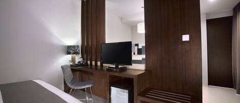 A modern hotel room with a bed, a desk with a television, a chair, and a mini-fridge.