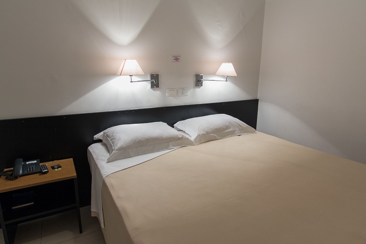 Ett hotellrum med en säng, två sänglampor, ett nattduksbord med en telefon och en fjärrkontroll samt en skylt på väggen.