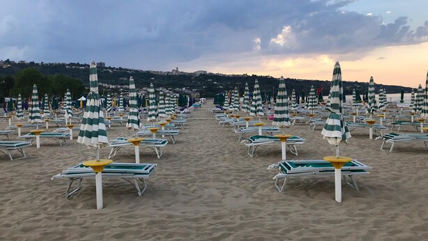 A beach with rows of sun loungers and striped umbrellas, a sandy path leading to the water, and a distant view of buildings on a hill. - Hotel Royal (Vasto)