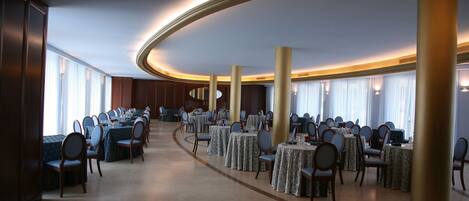 A spacious banquet hall with multiple round tables and chairs, large columns, and a curved ceiling.
