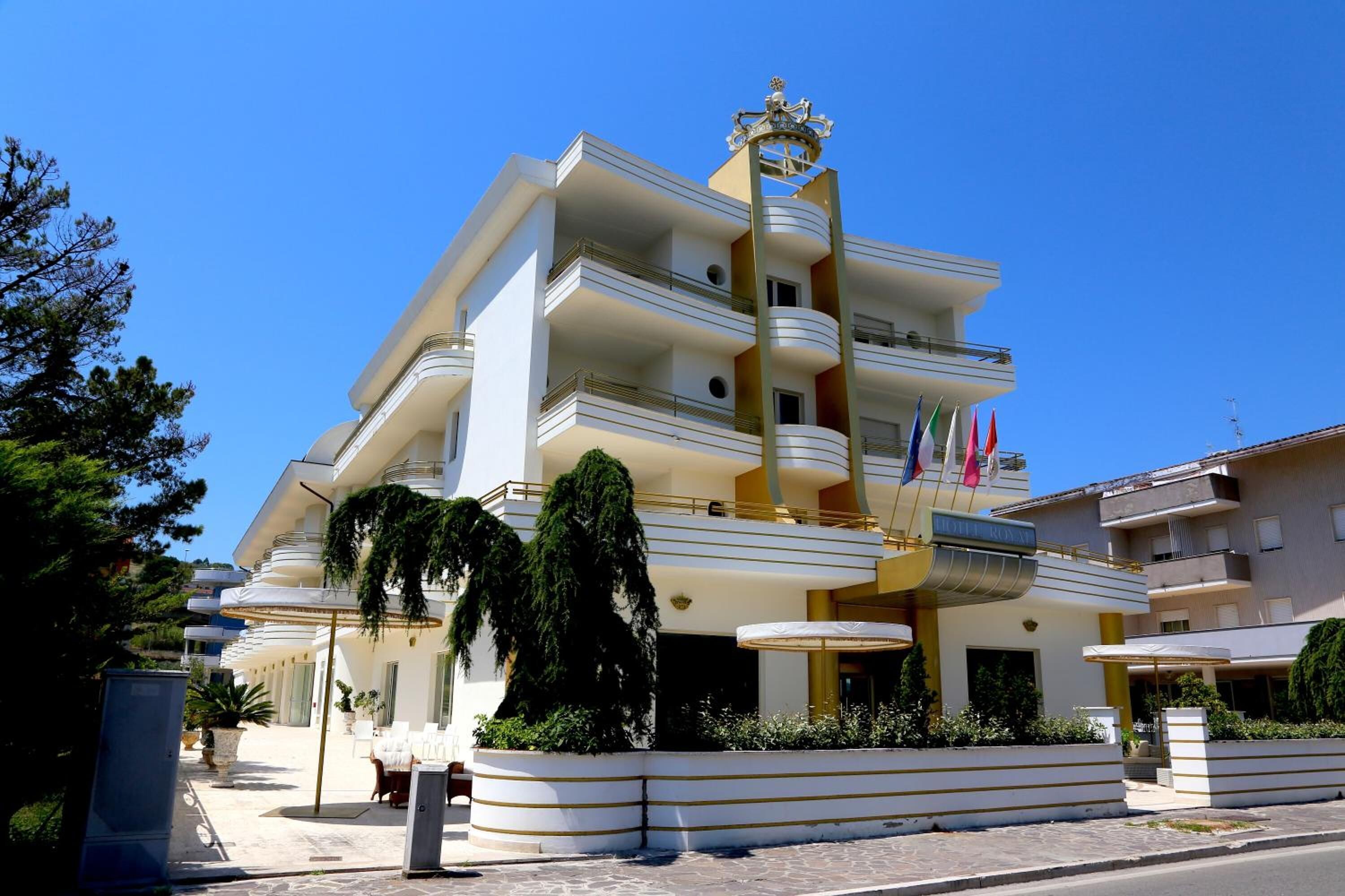 Un hotel a più piani con un balcone sporgente, un ingresso centrale e l'esposizione di una bandiera.