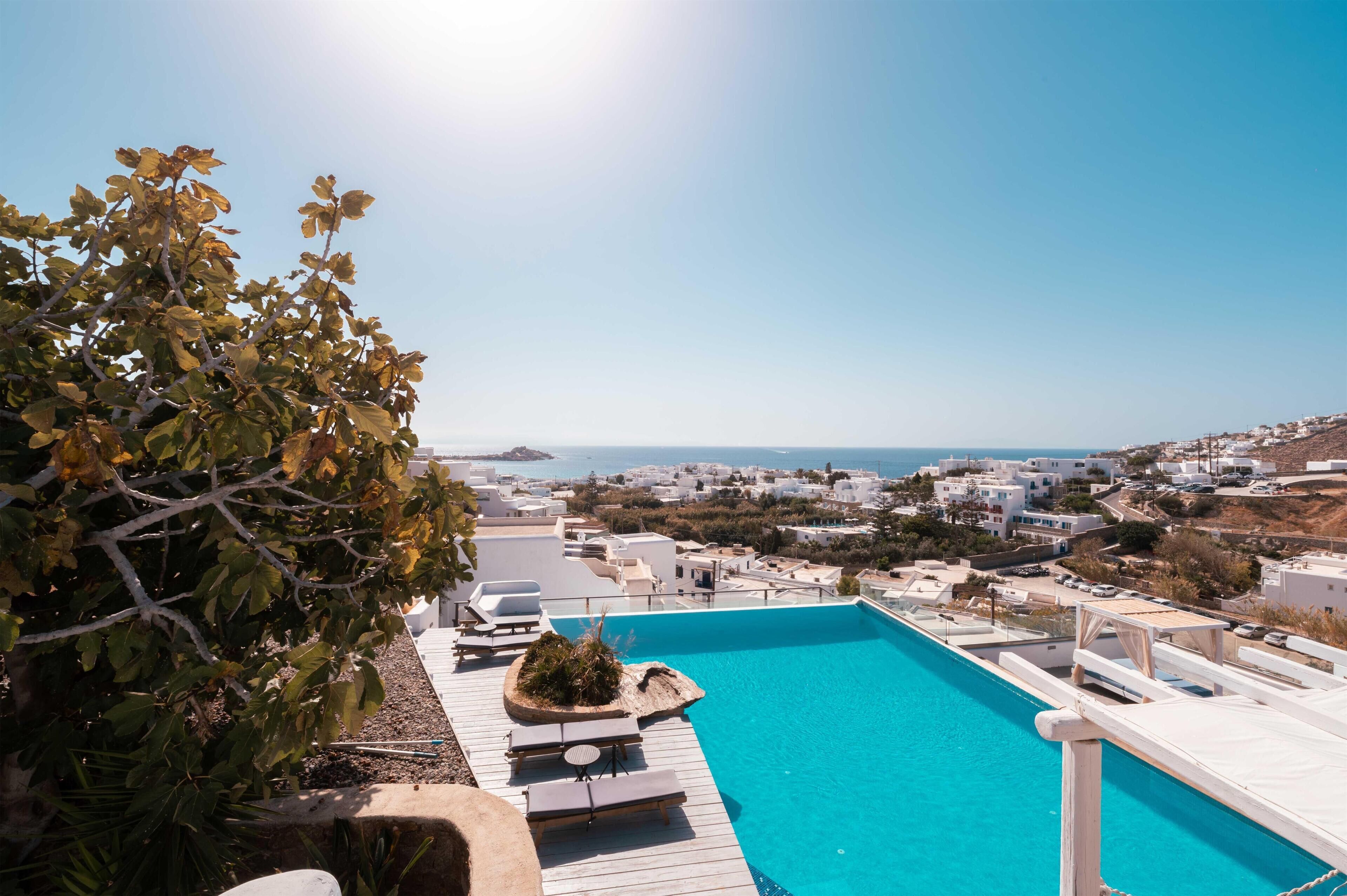 A clear blue pool with a wooden deck, surrounded by white buildings and a view of the sea.