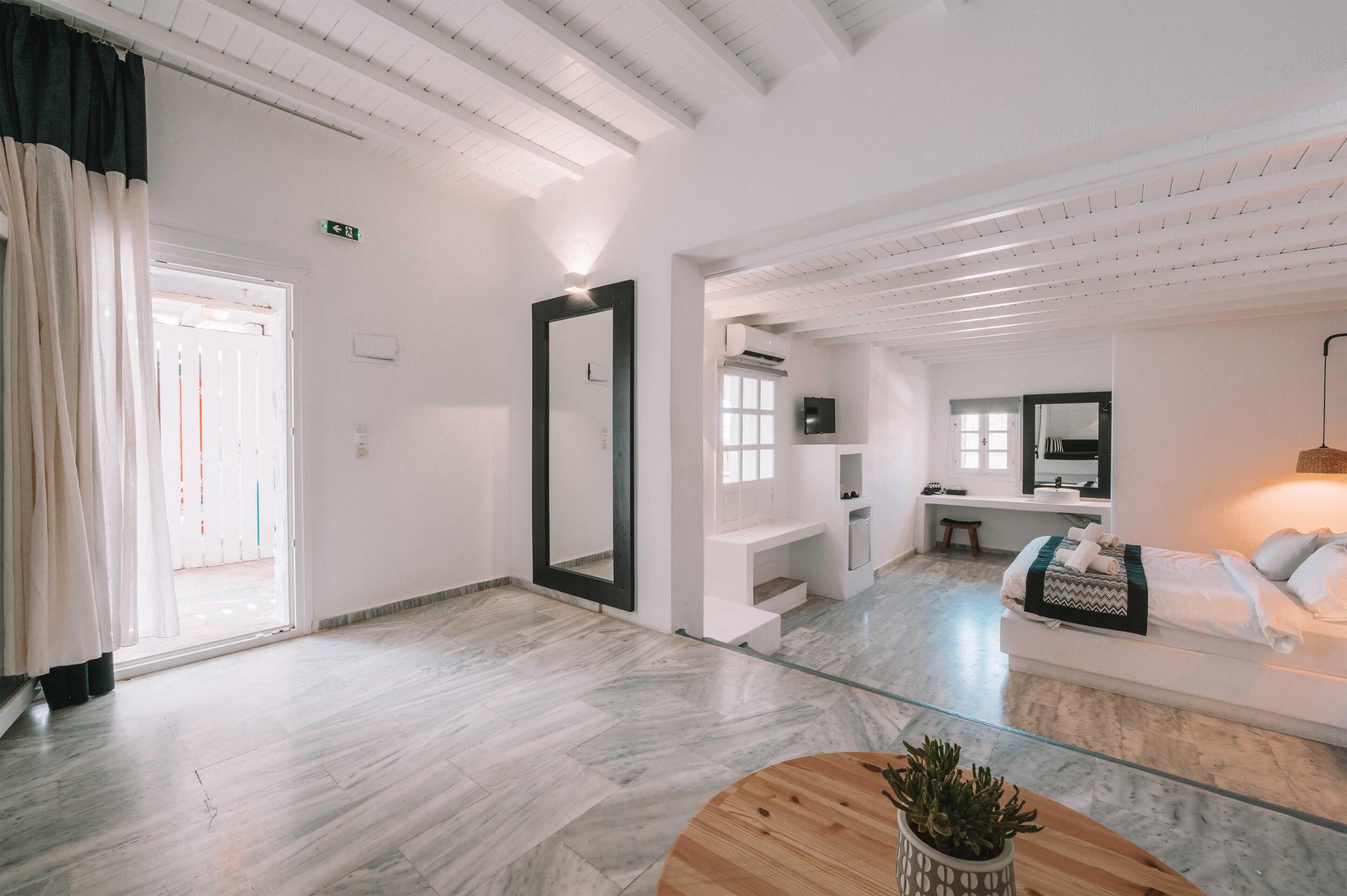 Una habitación de hotel moderna con una cama grande, una zona de estar con sofá y mesa, un espejo y una pequeña kitchenette.
