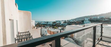 Una terraza en la azotea con barandilla de vidrio, edificios blancos al fondo y un cielo azul despejado.