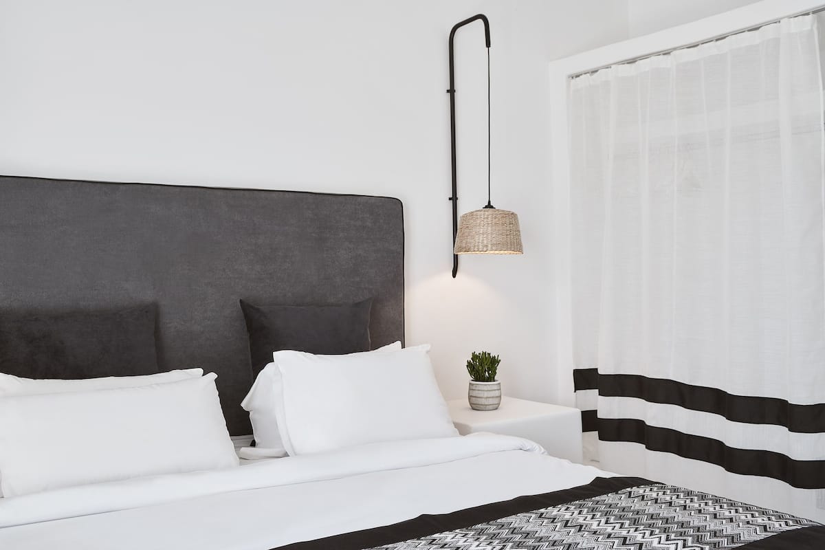 Une chambre moderne avec une tête de lit grise, du linge de lit blanc, une lampe de chevet, une petite plante en pot et un rideau à rayures noires et blanches.